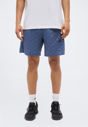 Mężczyzna noszący niebieskie spodenki Adidas z wzorem logo, białe skarpetki Adidas, czarne buty sportowe Adidas i prostą białą koszulkę, stojący na białym tle.