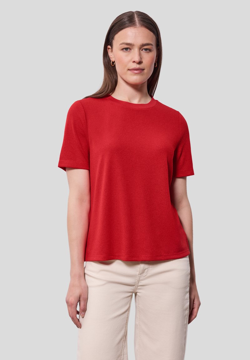 Červené tričko s krátkým rukávem vyrobené z jemného materiálu, s kulatým výstřihem a volným střihem. Vybaveno s kalhotami světlé barvy.