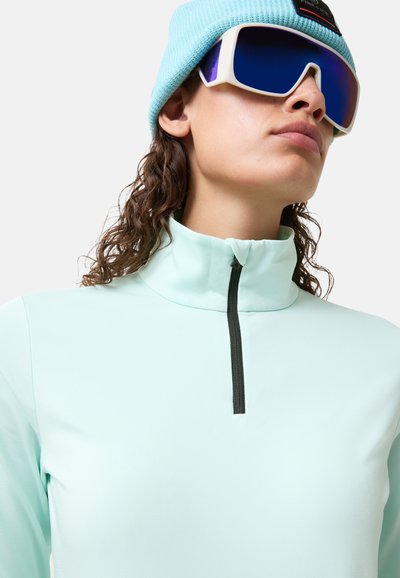 Hellblau, langärmeliger Pullover mit hohem Kragen und einem schwarzen Reißverschluss. Das Modell trägt eine hellblaue Strickmütze und große, reflektierende Sonnenbrillen.