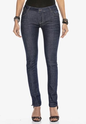 Kvinde iført mørkeblå slanke jeans og sorte højhælede sandaler med nitter, stående med afslappede hænder ned langs siderne.