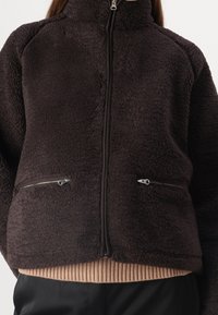 Giacca in pile marrone con colletto alto, chiusura lampo frontale e due tasche con zip. Accentuata da un orlo beige a coste. Texture morbida.