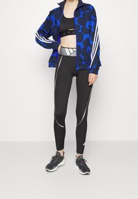 Chaqueta con cremallera de patrón azul y negro con rayas blancas, sujetador deportivo negro y leggings negros con acentos reflectantes; combinada con zapatillas negras.