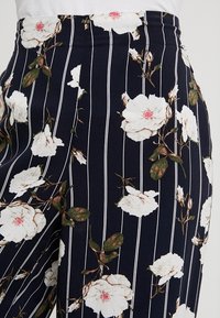 Marina högmidjade byxor med vita vertikala ränder, prydda med stora vita blommönster och gröna inslag, tillverkade av mjukt tyg.