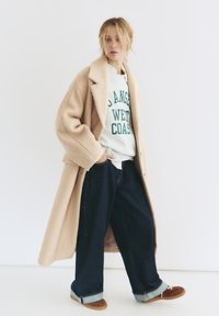 Beżowy wełniany płaszcz noszony na szarym swetrze z zielonym nadrukiem, w zestawie z ciemnoniebieskimi dżinsami o szerokich nogawkach i brązowymi zamszowymi butami.