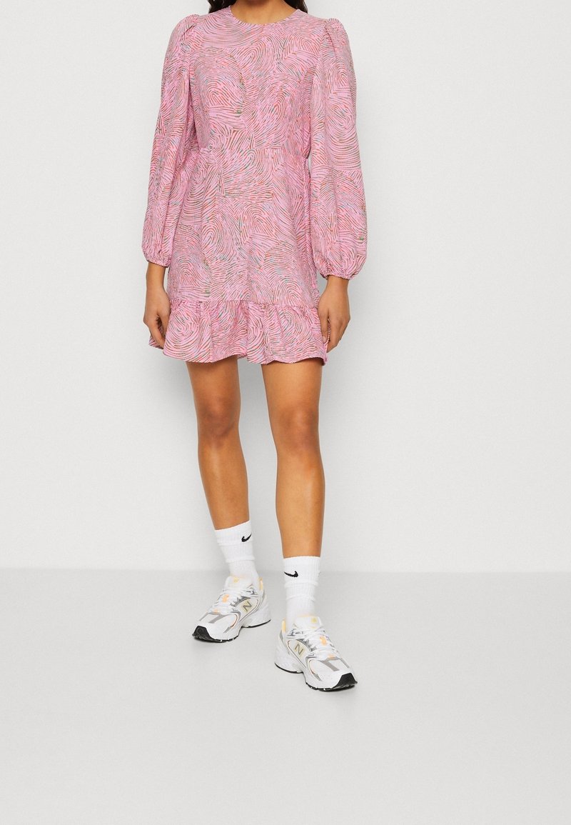 Robe rose à motifs avec manches longues bouffantes, ourlet à volants et design texturé. Associée à des baskets blanches et des chaussettes basses à logo.