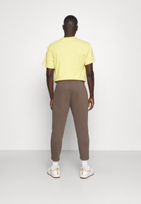 Gul bomullströja och bruna sweatpants, båda åtsittande, med vita sneakers med guldaccenter. Utsikt bakifrån.