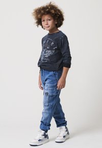 Fekete, hosszú ujjú póló grafikus nyomattal, világoskék cargo nadrággal, amely több zsebbel rendelkezik, és szürke árnyalatokkal díszített fehér sportcipővel párosítva.