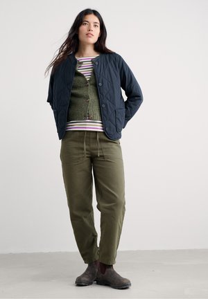Gesteppte marineblaue Jacke mit Knöpfen, darüber ein grüner Pullover und ein gestreiftes Hemd getragen. Olivgrüne Hose und braune Stiefeletten vervollständigen das Outfit.