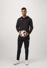 Schwarze sportliche Kapuzenjacke mit drei weißen Streifen, kombiniert mit schwarzen Hosen mit blauen Akzenten. Hält einen schwarz-weißen Fußball.