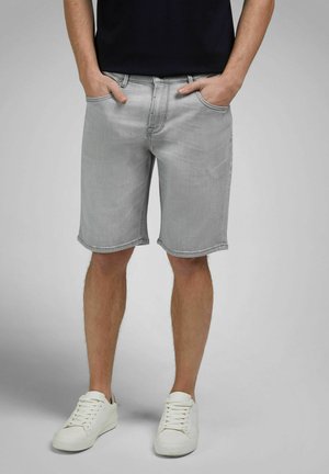 Man in lichtgrijze denimshorts met handen in de zakken en witte sneakers, staand tegen een effen achtergrond.