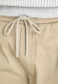 Pantalones de tiro ajustable en color beige con una textura suave, acompañados de un suéter de punto gris claro en la parte superior y cordones con puntas metálicas para el ajuste.
