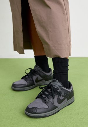 Personne portant des baskets Nike noires et grises avec des chaussettes noires et un pantalon beige court, debout sur un sol vert.