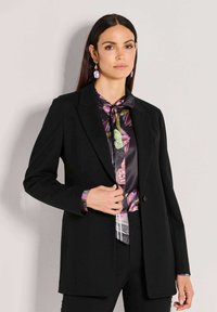 Frau mit glattem, dunklem Haar trägt ein schwarzes Blazer über einer Seidenbluse mit Schmetterlingsmuster und passenden Ohrringen, steht vor einer einfachen Wand.