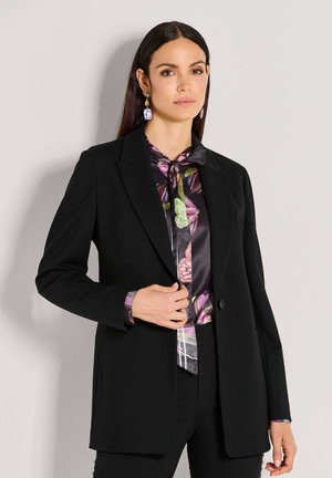 Woman with straight dark hair wears black blazer over a butterfly-patterned silk blouse and matching earrings, standing against a plain wall.