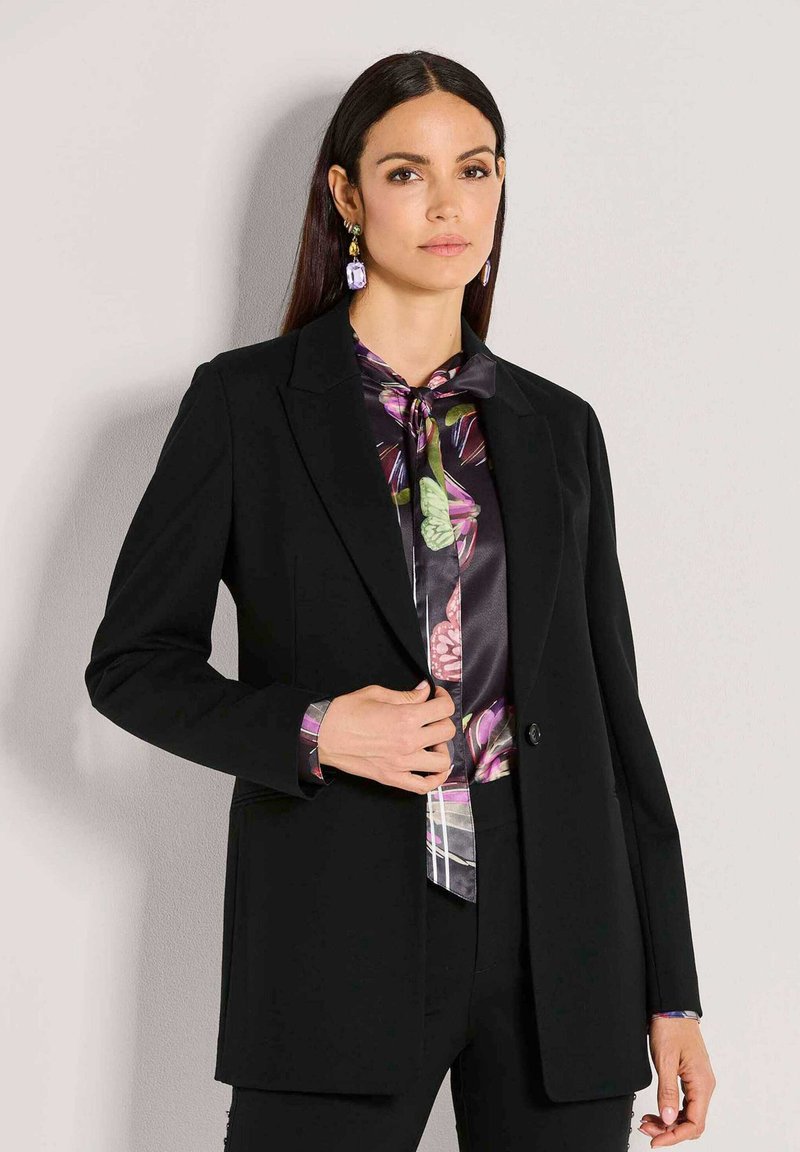Frau mit glattem, dunklem Haar trägt ein schwarzes Blazer über einer Seidenbluse mit Schmetterlingsmuster und passenden Ohrringen, steht vor einer einfachen Wand.