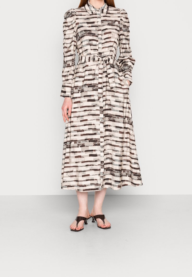 Femme portant une robe midi à manches longues ceinturée avec un motif abstrait en briques noir et blanc et des sandales à talons ouvertes noires.