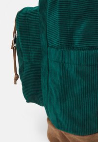 Green corduroy backpack with vertical ribbed texture and brown suede bottom. Features a zippered side pocket and decorative leather accents.