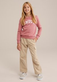 Gaišs rozā džemperis ar uzrakstu "AMOUR", brūnas krāsas cargo bikses ar lieliem sānu kabatām un pelēki sporta apavi ar baltām zolēm. Neitrāla fonā.