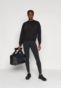 Czarny sweter z logo "DKNY", połączony z czarnymi legginsami i trampkami. Na zdjęciu znajduje się również czarna torba podróżna z akcentem w postaci kokardy. Gładkie tło.