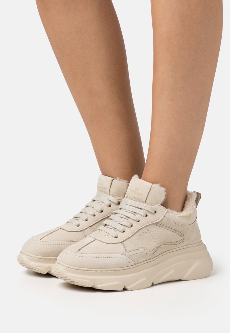 Baskets beiges à lacets avec semelles épaisses et doublure en fourrure portées sur des jambes nues sur un fond clair uni.