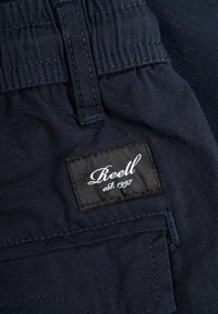 Tela de color azul marino con una textura suave; presenta una etiqueta cosida que dice "Reell" en escritura blanca y "est. 1997" sobre un fondo negro.