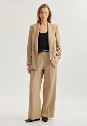 Beige skræddersyet blazer med vide bukser, parret med en sort tanktop. Stoffet ser glat ud; har et struktureret design og et bælte.
