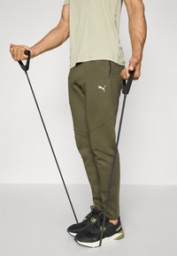 Olivgrüne Sweatpants mit Seitentaschen und schwarzen Akzenten, kombiniert mit schwarzen Sportschuhen und Halteband.