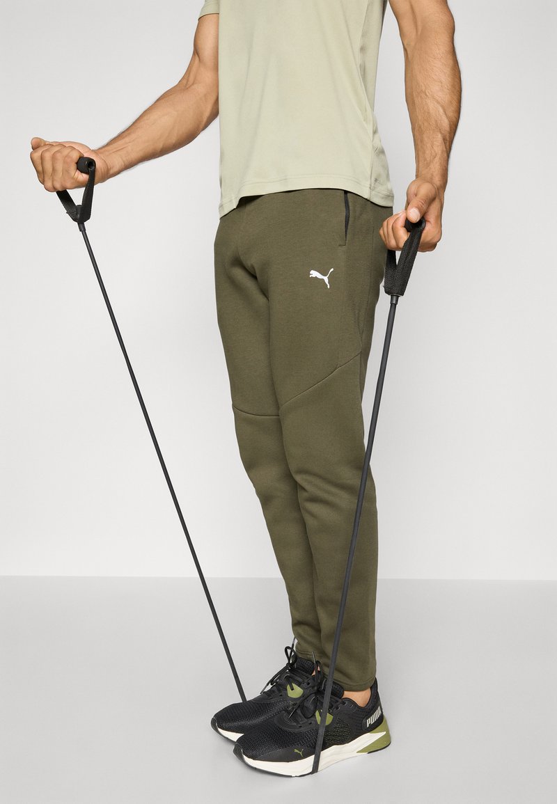 Pantalons de survêtement vert olive avec poches latérales et accents noirs, associés à des chaussures de sport noires, tenant des bandes de résistance.