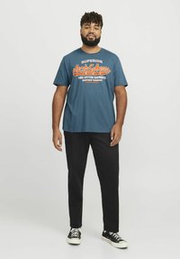 Camiseta gráfica de algodón azul con el logo "Jack & Jones" en naranja y blanco, combinada con pantalones negros y zapatillas negras con cordones blancos.