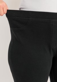 Zwarte stretchbroeken met een brede tailleband, voorzien van een enkele voorzak. Het materiaal heeft een soepele, gestructureerde afwerking.