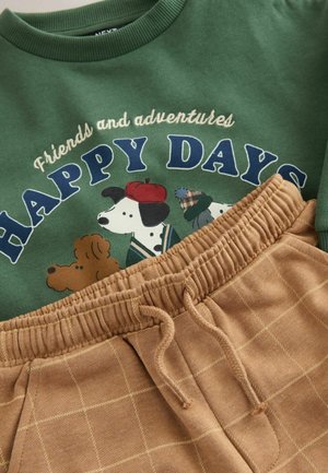 Grøn T-shirt med hundegrafik og teksten "HAPPY DAYS", parret med tan sweatpants med gittermønster og elastisk talje.