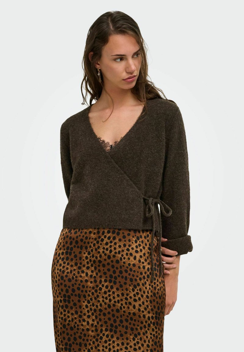 Femme portant un pull cache-cœur marron foncé avec un nœud sur le côté et une jupe imprimée léopard, regardant vers sa gauche sur un fond uni.