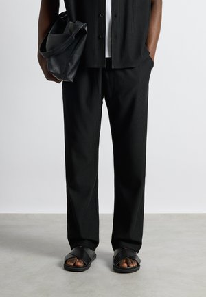 Personne portant un pantalon noir à rayures fines et des sandales noires, tenant un sac en cuir noir dans une main, l'autre main dans la poche.