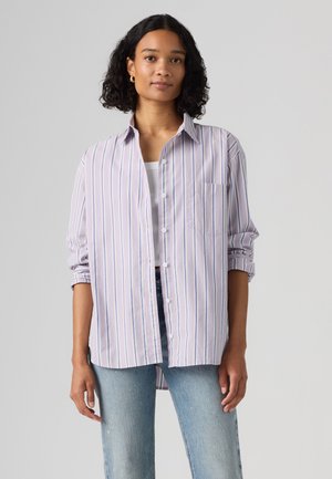 Femme aux cheveux bouclés portant une chemise boutonnée blanche et violet clair à rayures verticales par-dessus un haut blanc et un jean bleu, sur un fond uni.