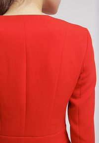 Femme portant un blazer rouge ajusté, vue de dos, avec de longs cheveux bruns reposant sur son cou.