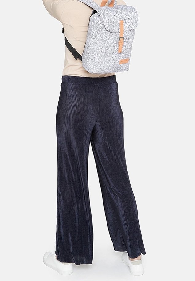 Personne portant un pantalon large bleu marine, des baskets blanches, un haut beige, et portant un sac à dos gris à motif avec des détails de sangle couleur camel.