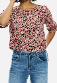 Kobieta w bluzce w kwiaty w kolorze różowym i beżowym z rękawami do łokcia, włożonej do niebieskich dżinsów.