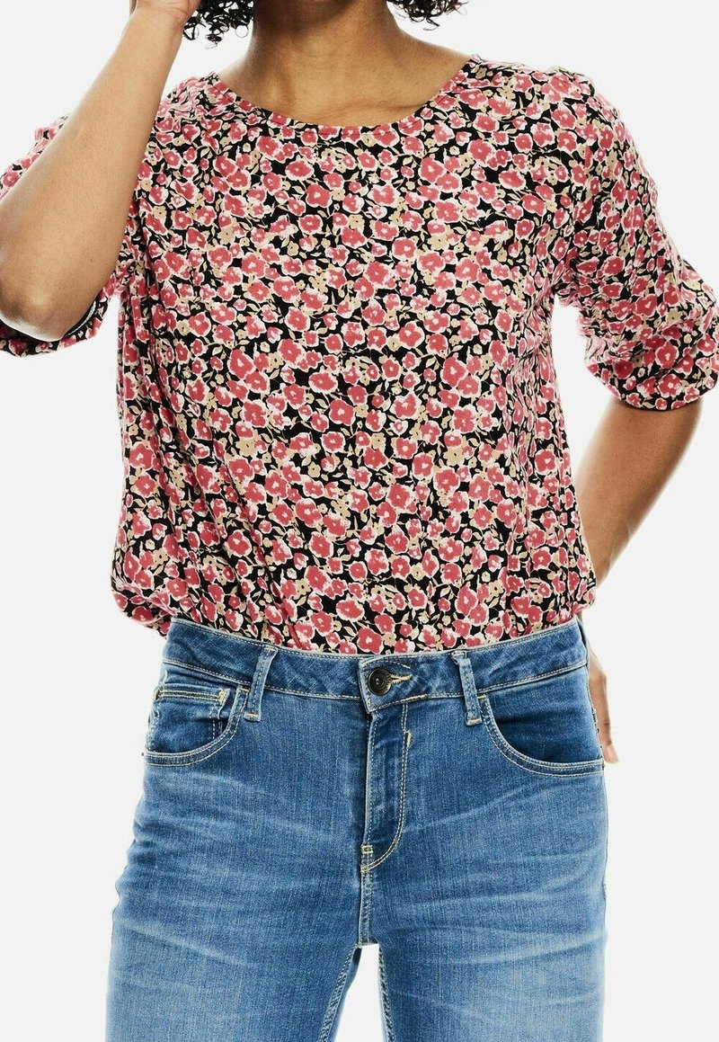 Kobieta w bluzce w kwiaty w kolorze różowym i beżowym z rękawami do łokcia, włożonej do niebieskich dżinsów.