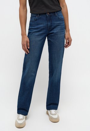 Personne portant un jean bleu foncé droit, des baskets blanches et beiges, et un haut noir à manches courtes avec les bras détendus le long du corps.