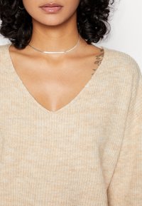Pull beige en tricot avec un col en V, présentant une texture douce et un motif subtil. Comprend un collier argenté et un tatouage visible.