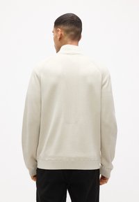 Άνδρας που φοράει ελαφρύ μπεζ πλεκτό πουλόβερ με υφασματιστές επιφάνειες, γυρισμένος με την πλάτη προς τα έξω, σε απλό λευκό φόντο.