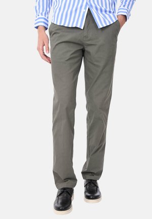 Uomo che indossa pantaloni verde oliva a gamba dritta, scarpe nere con lacci e una camicia a righe blu e bianca con le mani in tasca.