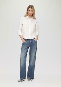 Chemise blanche à boutons avec de petits motifs de cœurs roses, associée à un jean en denim bleu clair à jambes larges et des chaussures argentées. Manches légèrement repliées.