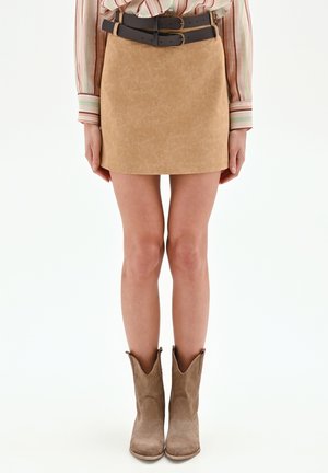 Donna che indossa una minigonna in camoscio beige con due cinture nere, una camicia a righe a maniche lunghe e stivali da cowboy in camoscio color cuoio, in piedi dritta.