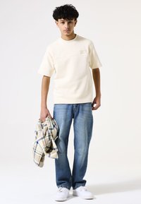 Crème t-shirt met korte mouwen en een gestructureerde voorkant, gecombineerd met lichtblauwe jeans. Houdt een geruite flanellen overhemd vast in beige, blauw en groen.