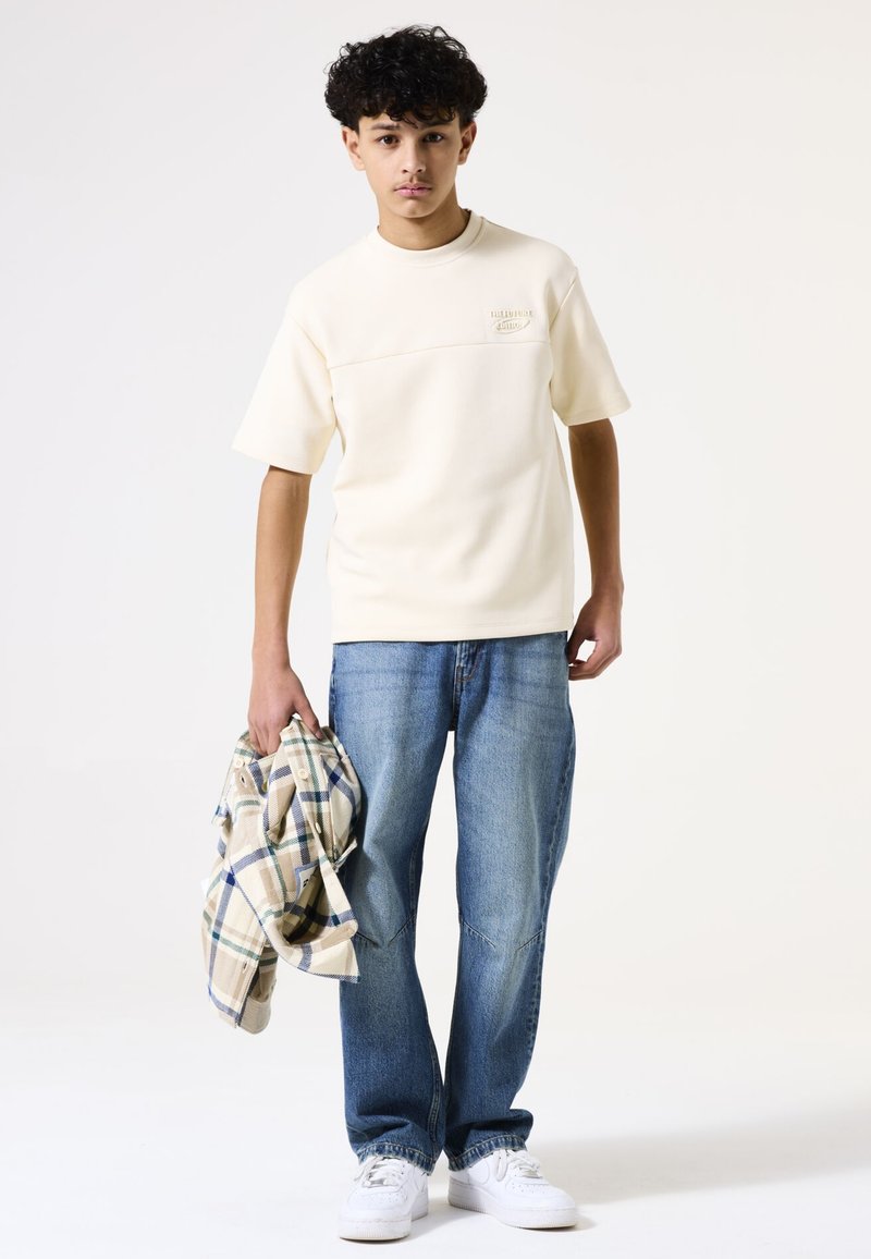 Crème t-shirt met korte mouwen en een gestructureerde voorkant, gecombineerd met lichtblauwe jeans. Houdt een geruite flanellen overhemd vast in beige, blauw en groen.