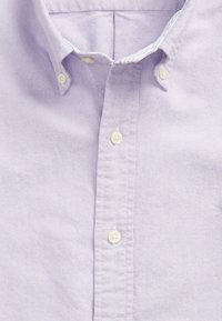 Svetle fialová košile s klasickým límcem, která má bílé knoflíky a texturovanou látku. Podrobné šití podél lég. 