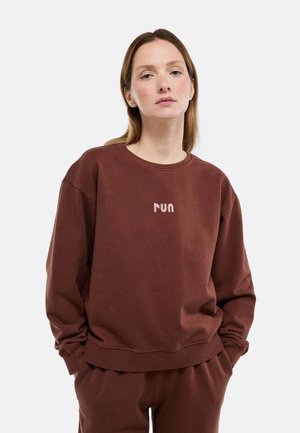 Femme portant un sweat-shirt marron et un pantalon, les mains dans les poches, le sweat-shirt comporte un petit texte brodé « run » au centre devant.