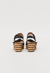 Sorte skinnwedge sandaler med korkhæl, med et lagdelt design og en spenne-stropp. Bakre visning viser en buet, teksturert hæl.