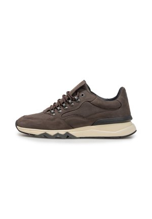Dunkelbrauner Sneaker mit beiger Sohle, Metallösen und Zickzack-Profil, seitlich auf weißem Hintergrund gezeigt.